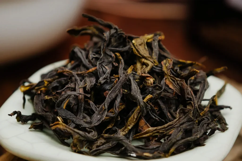 A mound of dark, twisted loose-leaf tea with golden-brown tips rests on a white plate, photographed close-up in warm, softly lit surroundings.