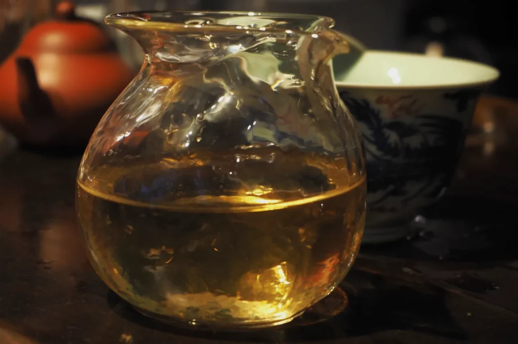 A glass pitcher of golden amber tea sits on a dark table; a patterned cup and orange teapot behind it create a warm, calm mood.