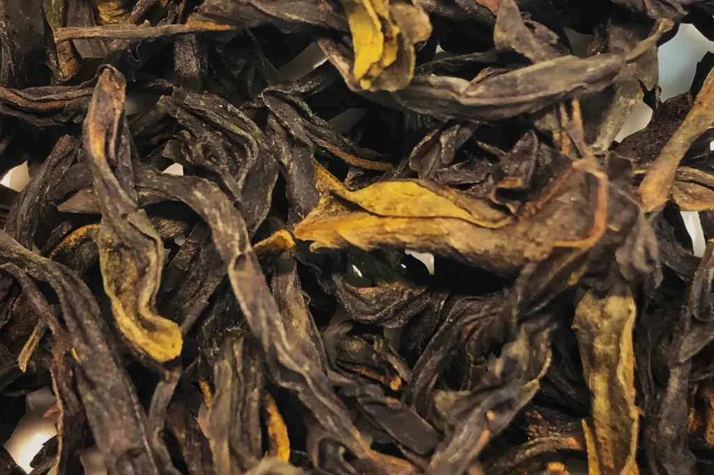 Close-up of dried tea leaves, dark brown and twisted with occasional golden-yellow edges, densely layered to create a rustic, earthy texture.