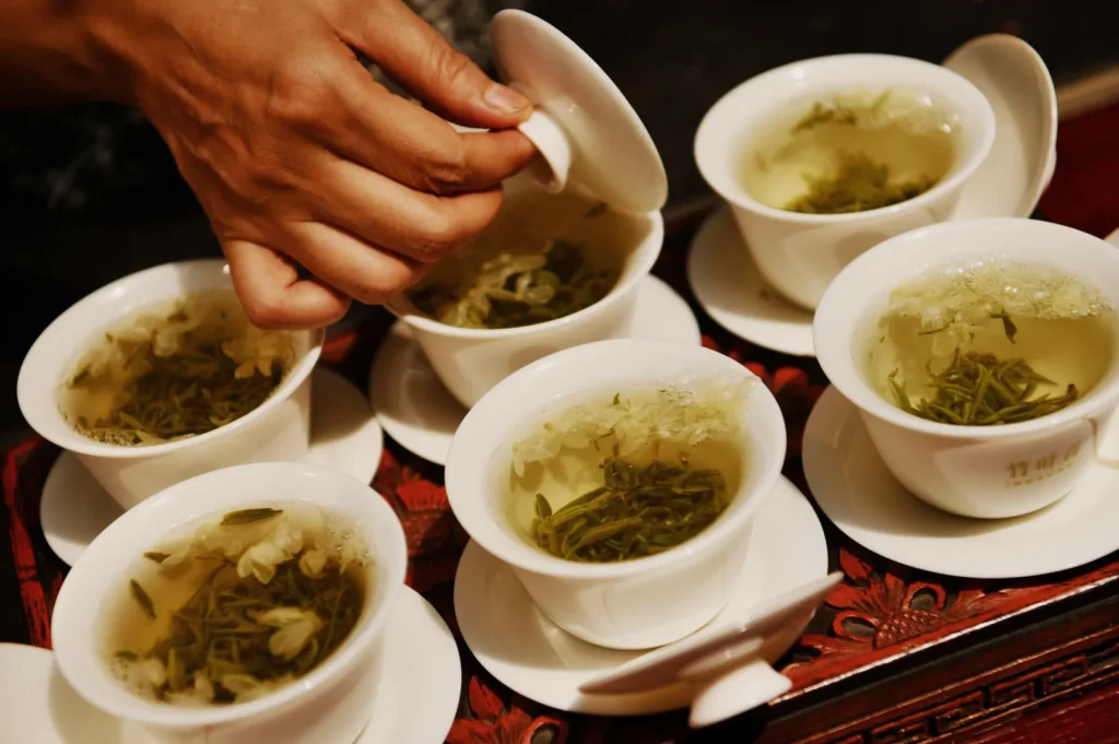 A hand lifts a lid from a white gaiwan as several cups of steeped green tea sit on a red patterned tray, creating a calm tea ceremony scene.