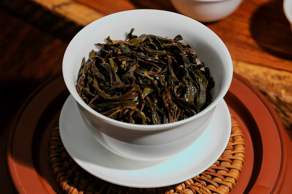 A white bowl filled with dark, twisted loose-leaf tea rests on a matching saucer atop a woven coaster and wooden tea tray, evoking a warm, calm tea setting.