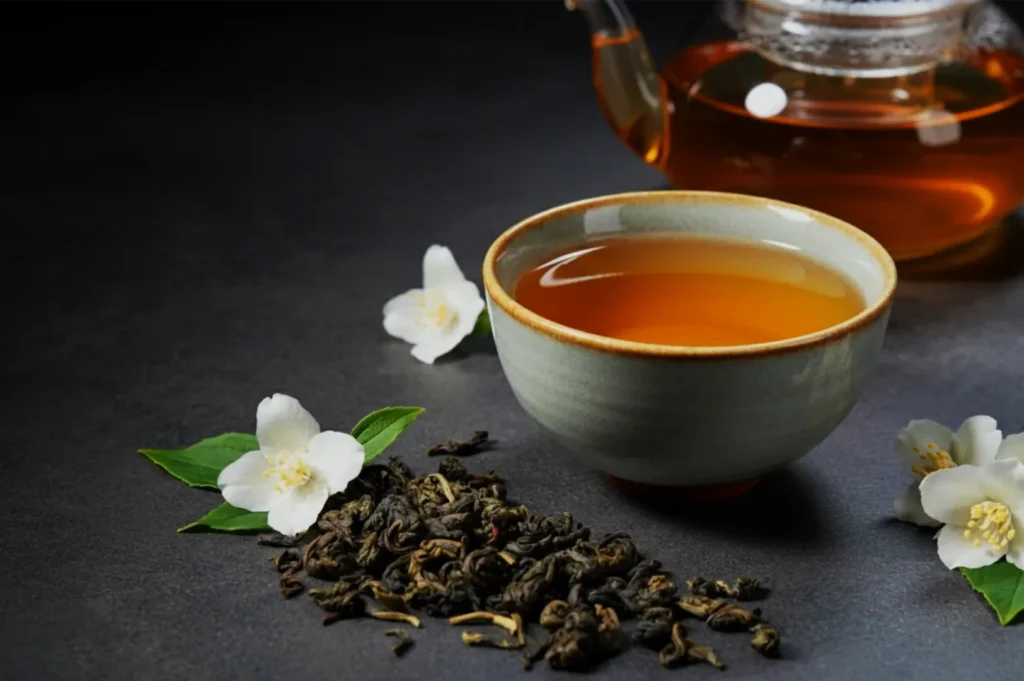 A ceramic cup of amber tea sits beside a glass teapot, loose tea leaves, and white blossoms on a dark slate surface, creating a calm, elegant mood.