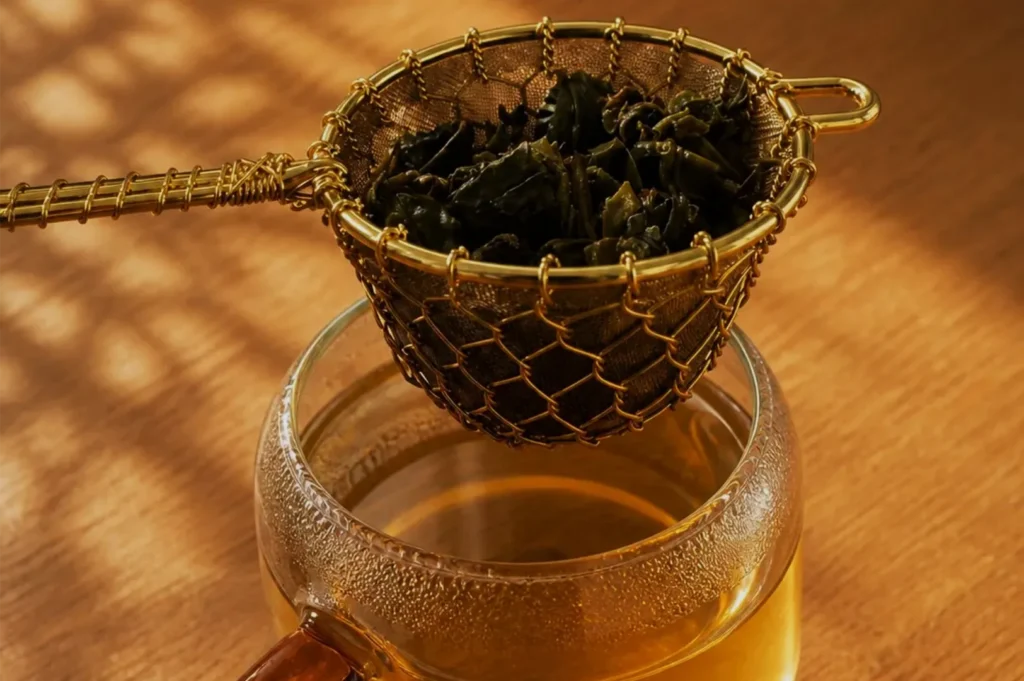 A brass mesh strainer filled with dark loose tea leaves rests over a glass teapot on a wooden surface, evoking a warm, quiet tea-making ritual.