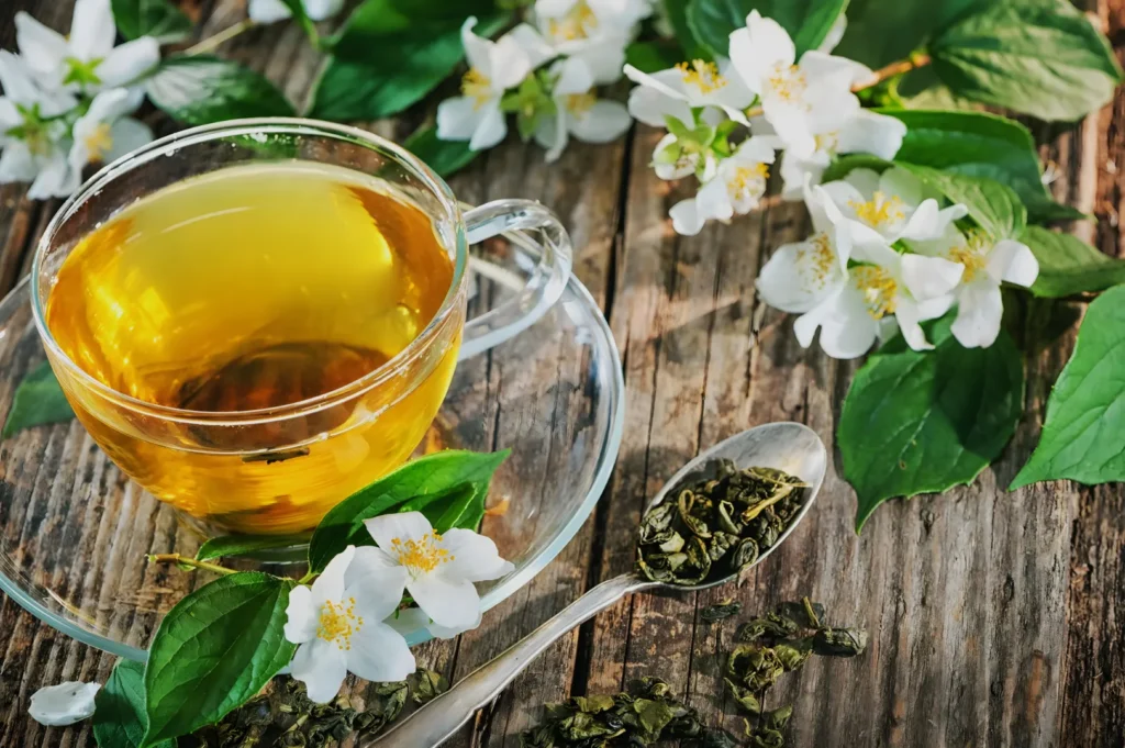 A glass cup of golden jasmine tea rests on a rustic wooden table beside loose tea leaves, a spoon, green foliage, and white jasmine blossoms, evoking calm.