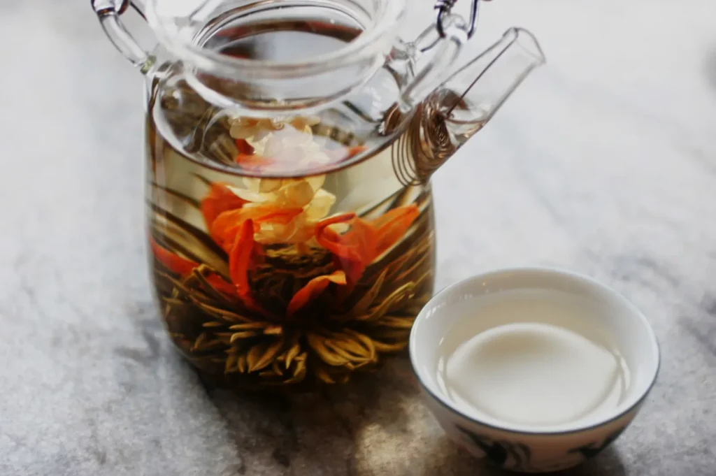 A glass teapot steeps orange and yellow flowers with herbs in clear tea, beside a small white cup on a stone surface, creating a calm, natural mood.