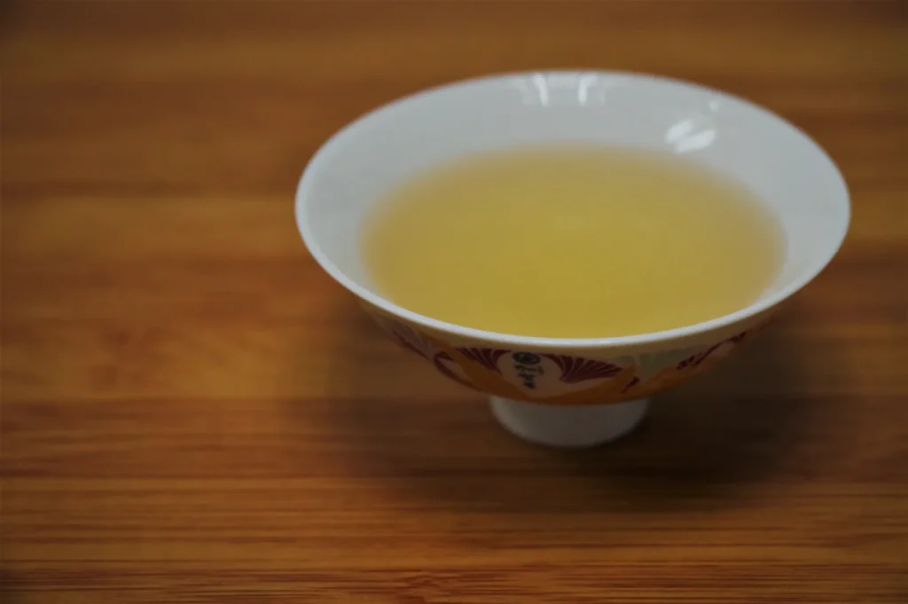 A white porcelain bowl filled with pale golden tea sits on a warm wooden table, creating a quiet, simple, inviting scene.