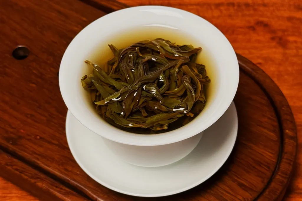 A white gaiwan holds brewed loose-leaf tea, with glossy green-brown leaves in amber liquor on a warm wooden tea tray, creating a calm, traditional mood.