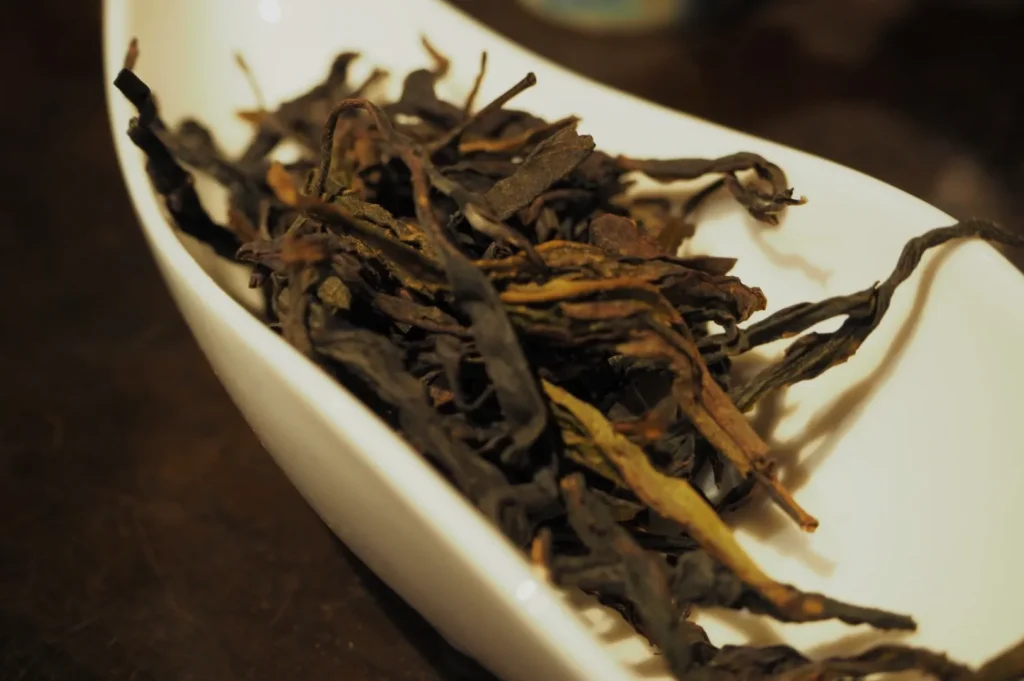 Close-up of dark, twisted dried tea leaves piled in a white dish on a wooden surface, creating a rustic, earthy and aromatic impression.