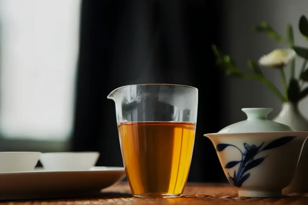 A clear glass of amber tea sits on a wooden table beside a blue-and-white ceramic teapot, with a softly lit plant in the background, creating a calm mood.
