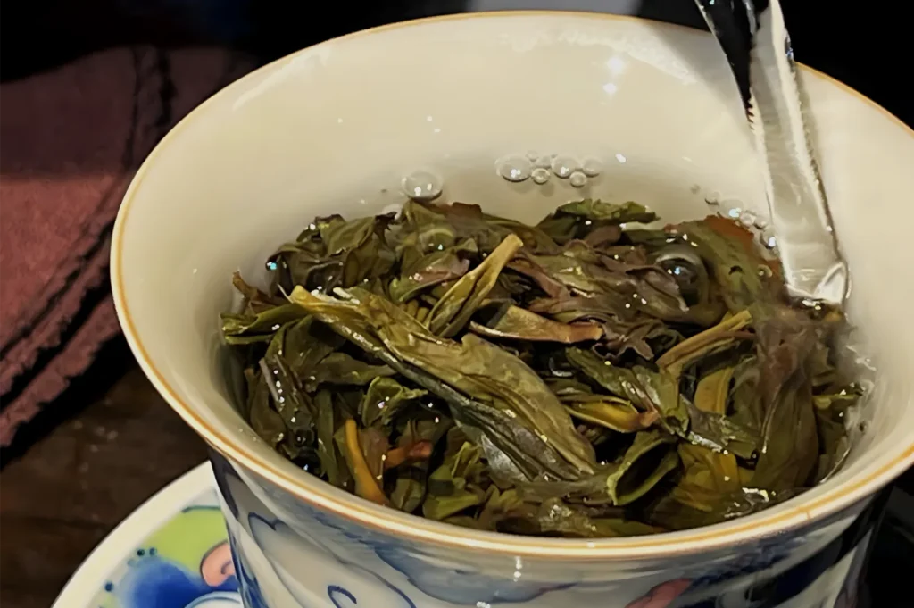 A white porcelain gaiwan holds brewed loose-leaf green tea, with wet leaves submerged in amber liquor and a lid resting at the rim. The scene feels calm and traditional.
