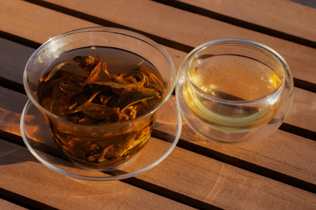 Two clear glass cups sit on a sunlit wooden table: one holds dark amber tea with loose leaves, the other pale golden tea, creating a warm, tranquil mood.