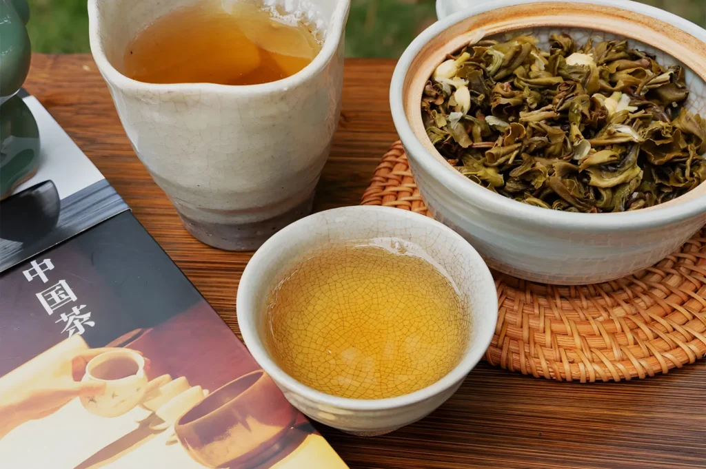 A calm Chinese tea setting: amber tea in a small cup beside a white pitcher and bowl of loose leaves, arranged on a wooden table with a tea book.