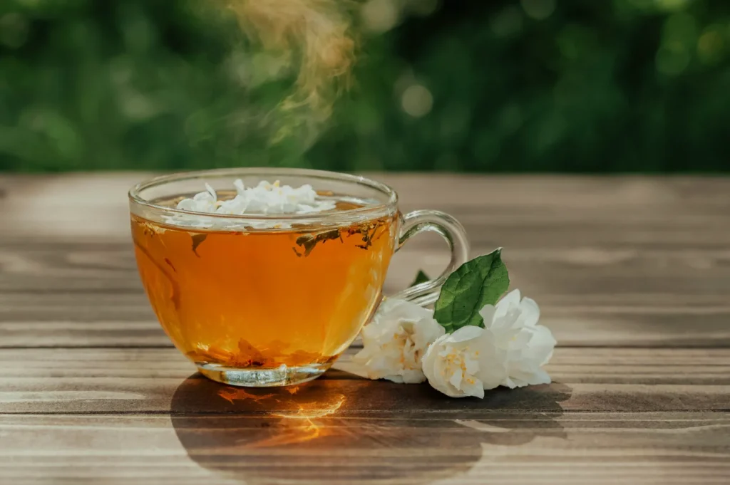 A steaming glass cup of amber tea rests on a wooden table beside white jasmine blossoms, with soft green foliage blurred in the calm background.