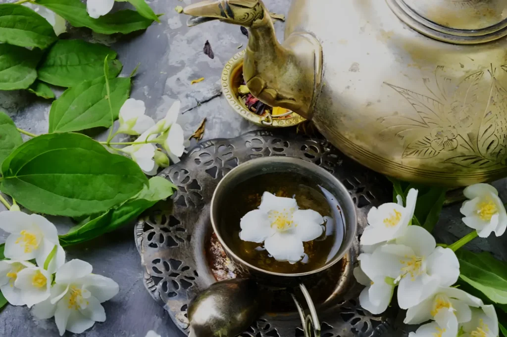 An ornate brass teapot pours tea into a small cup among white jasmine blossoms and green leaves, creating a tranquil, elegant still life.