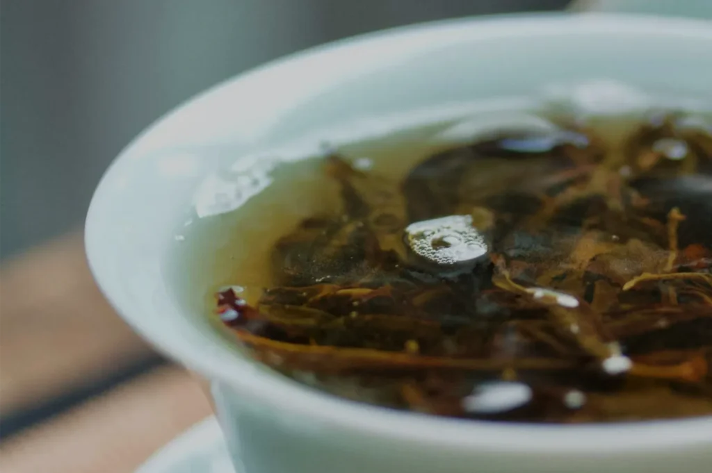 Close-up of dark herbal tea with floating leaves steeping in a white ceramic cup; tiny bubbles dot the surface, creating a calm, soothing mood.