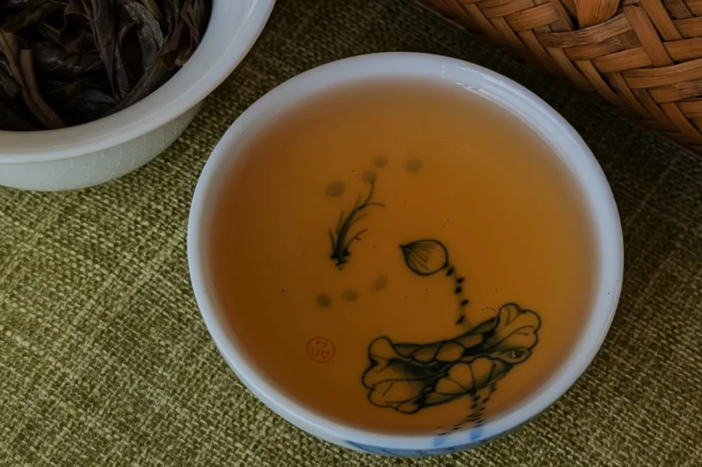 Overhead view of amber tea in a white porcelain cup decorated with a delicate plant and fish, beside dried tea leaves and woven baskets; calm, rustic mood.