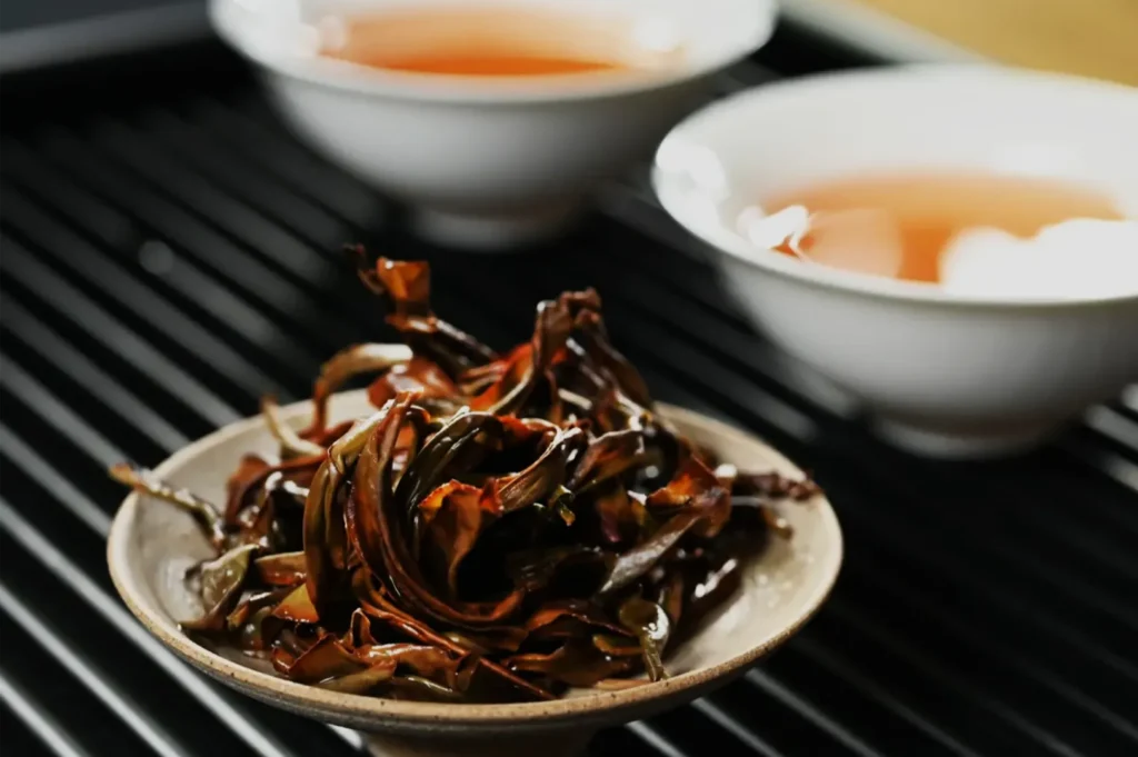 A ceramic plate piled with glossy, dark-brown curled strips sits on a black ridged surface, with two bowls of amber sauce behind it; moody food styling.