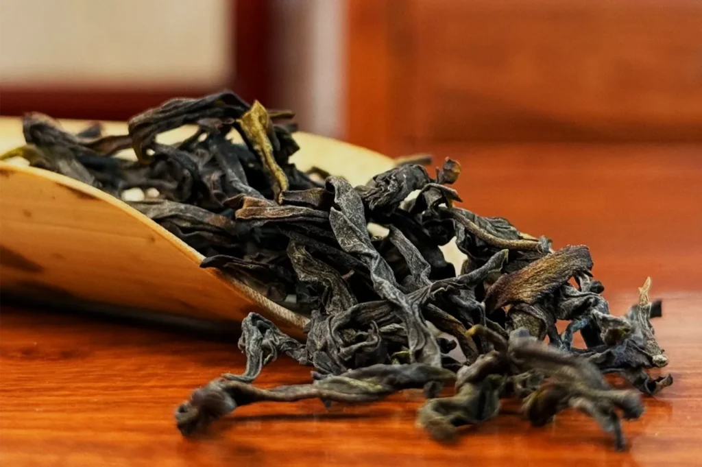 Close-up of loose, dark twisted tea leaves spilling from a shallow wooden scoop onto a polished table, creating a warm, rustic still life.