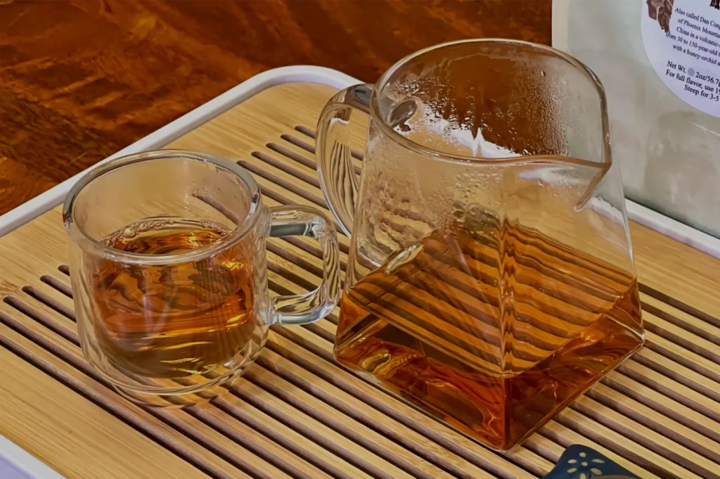 A glass cup of amber tea sits beside a larger glass pitcher on a bamboo tray, creating a warm, calm tea-serving scene.