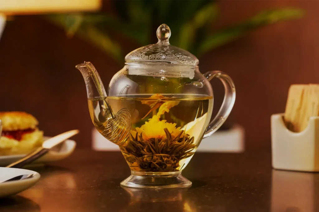 A clear glass teapot steeps blooming yellow tea flowers on a warm wooden table, with a cup and snacks nearby, creating a cozy, inviting mood.