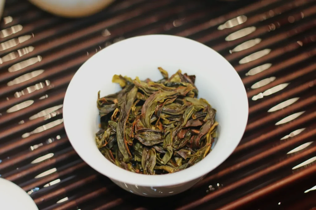 A white tasting cup filled with twisted, olive-green and brown loose-leaf tea rests on a dark wooden slatted tray, creating a calm, rustic tea-tasting scene.