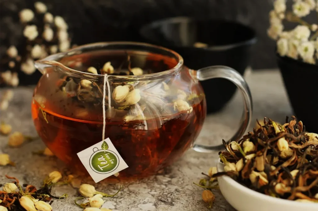 A glass teapot of amber herbal tea with a visible tea bag sits among dried flowers and herbs, creating a warm, rustic, calming scene.