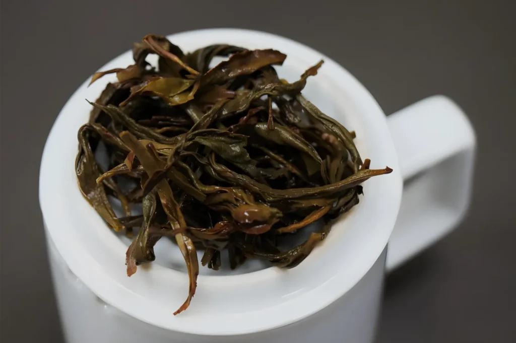A white mug holds a heap of dark, twisted loose tea leaves after brewing, shown against a plain gray background in a quiet, close-up view.