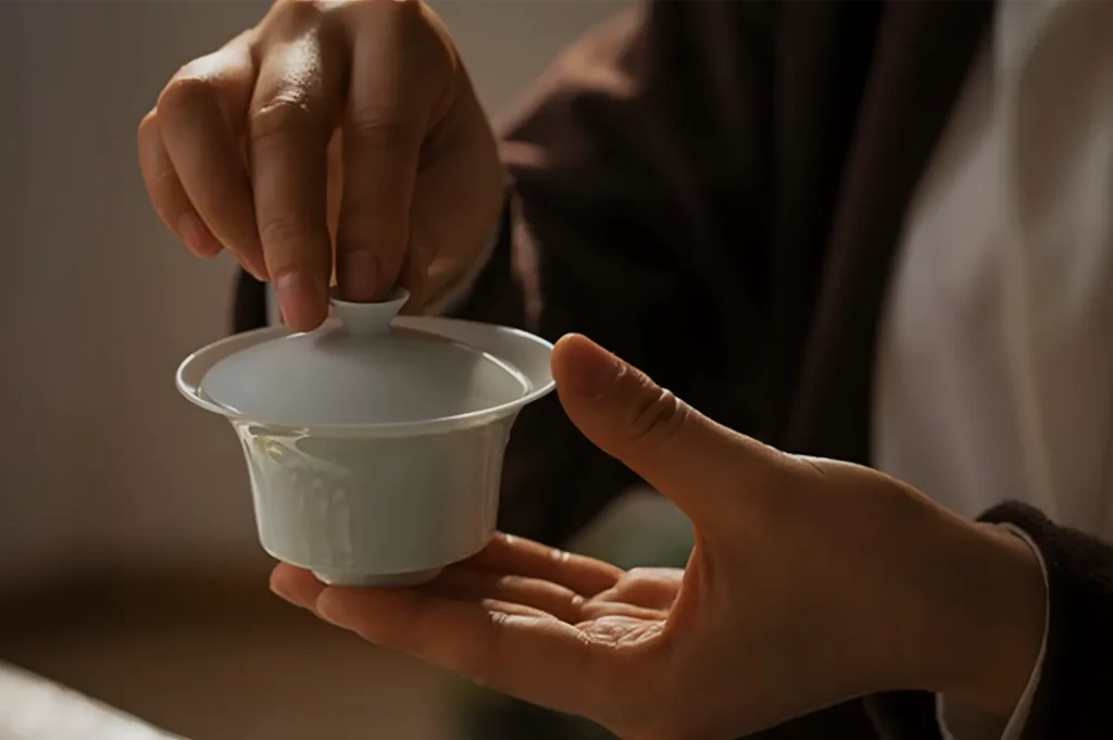 Close-up of hands carefully lifting a small white ceramic tea cup lid, revealing a traditional tea bowl; warm, soft lighting creates a calm, mindful mood.