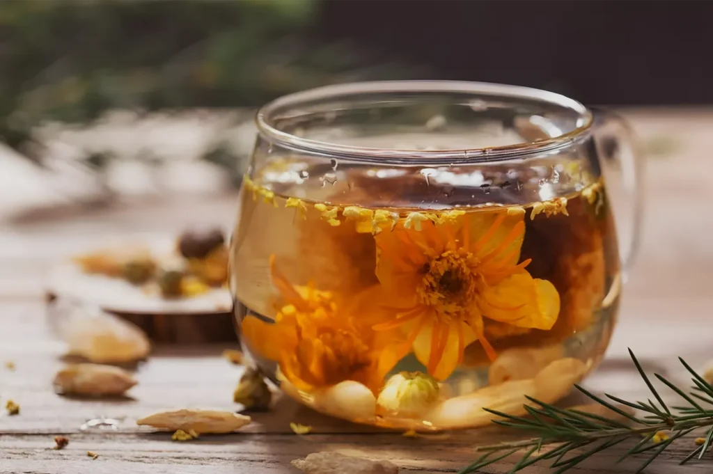 A glass cup of amber herbal tea steeping with bright orange calendula flowers sits on a rustic wooden table, creating a warm, calming mood.