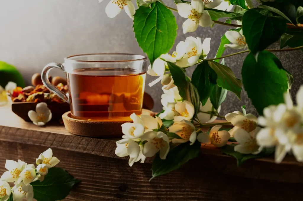 A glass cup of amber herbal tea sits beside dried botanicals and white blossoms with green leaves, creating a warm, calming natural still life.