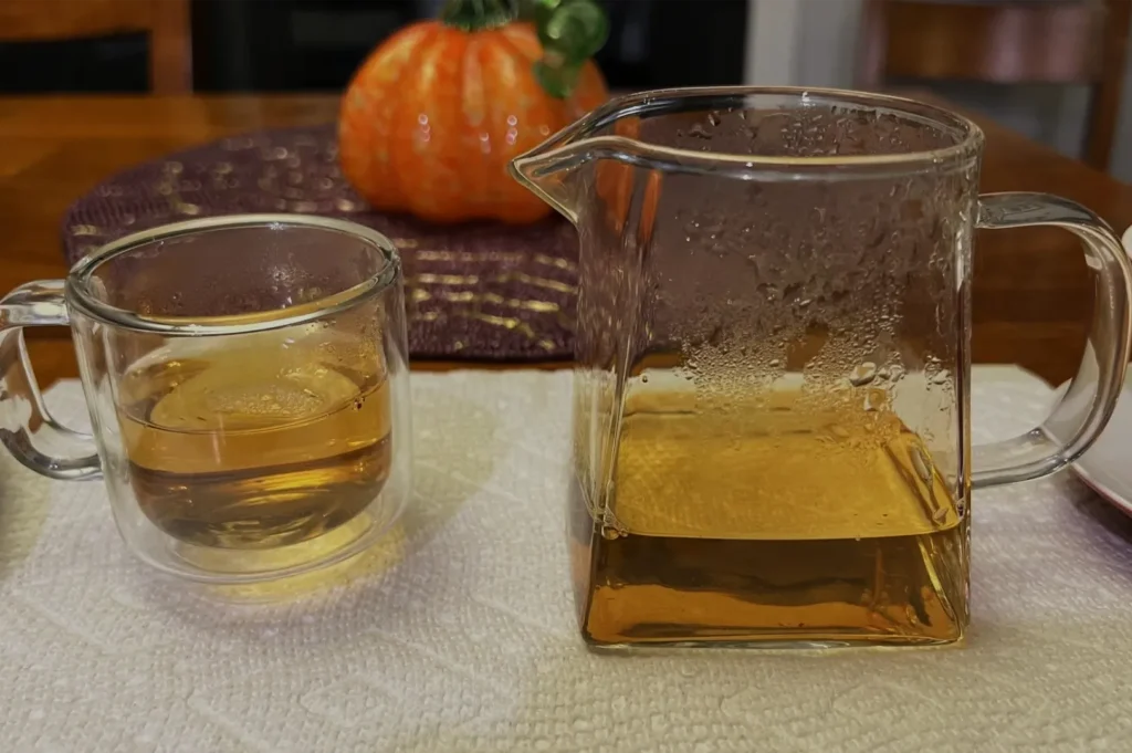 A clear glass cup and pitcher hold amber tea on a white cloth, with an orange pumpkin behind them, creating a cozy autumn table scene.