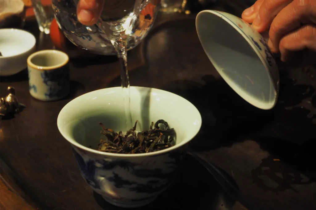 Hands pour water from a glass pitcher over loose tea in a blue-and-white porcelain gaiwan, its lid held aside in a calm tea ritual.