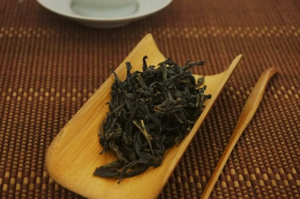 Loose dark tea leaves sit on a small bamboo scoop beside a wooden tea utensil, arranged on a woven brown mat in a calm, rustic tea setting.