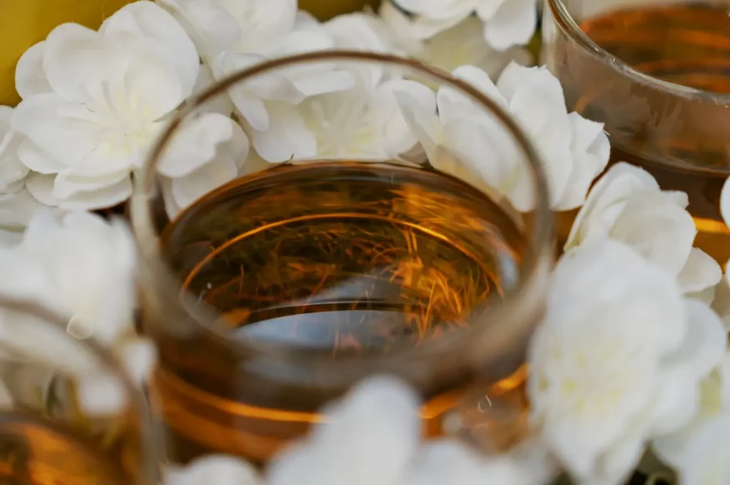 A glass cup of amber tea sits among white blossoms, with another cup in the background; soft light creates a calm, elegant mood.
