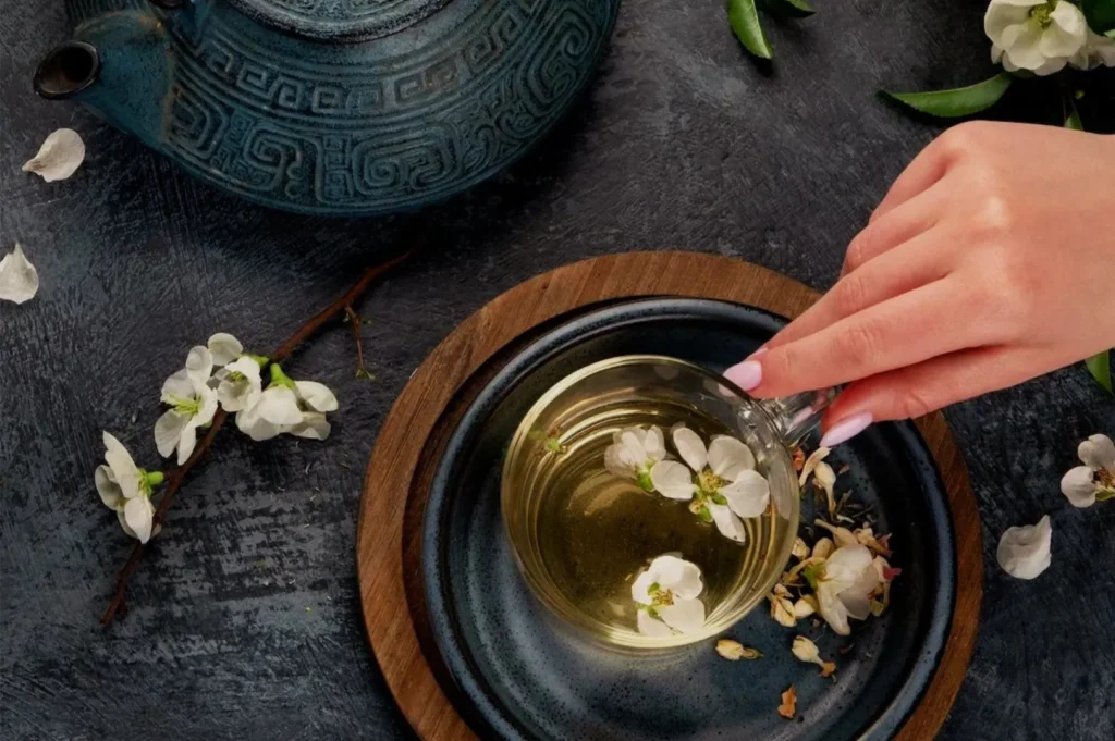 Overhead view of a hand reaching for herbal tea with floating white blossoms, beside a blue teapot and scattered flowers on dark stone.