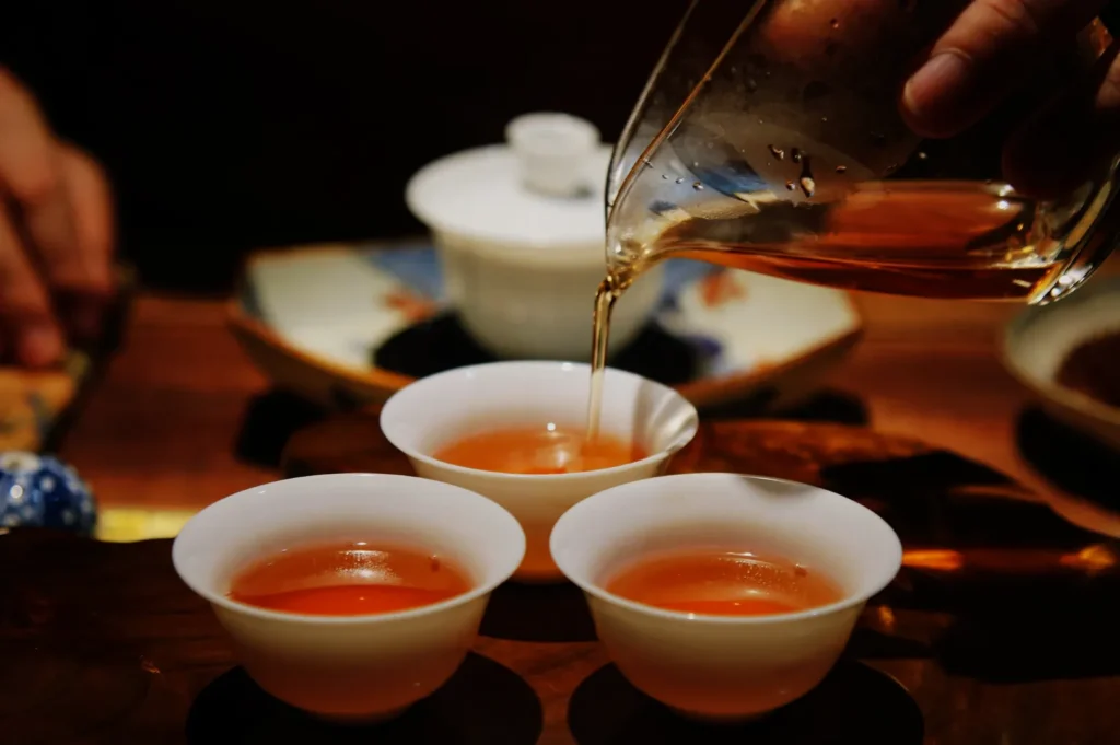 A hand pours amber tea from a glass pitcher into a small white cup, with two filled cups and a teapot on a wooden table, creating a warm, intimate mood.