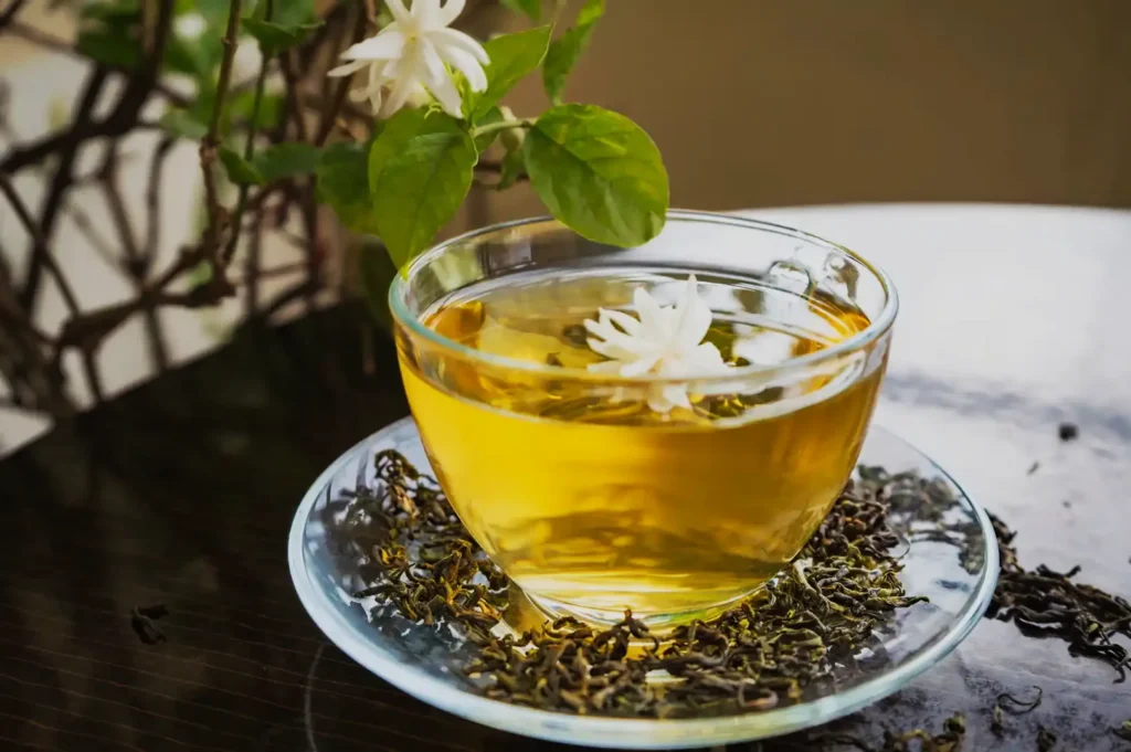 A clear glass cup of golden jasmine tea, topped with white blossoms, rests on a saucer of loose tea leaves beside fresh green foliage, creating a calm mood.
