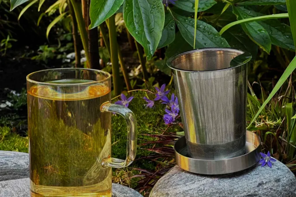 A glass mug of golden tea sits beside a stainless-steel tea infuser on garden stones, surrounded by lush leaves and purple flowers; calm, refreshing scene.