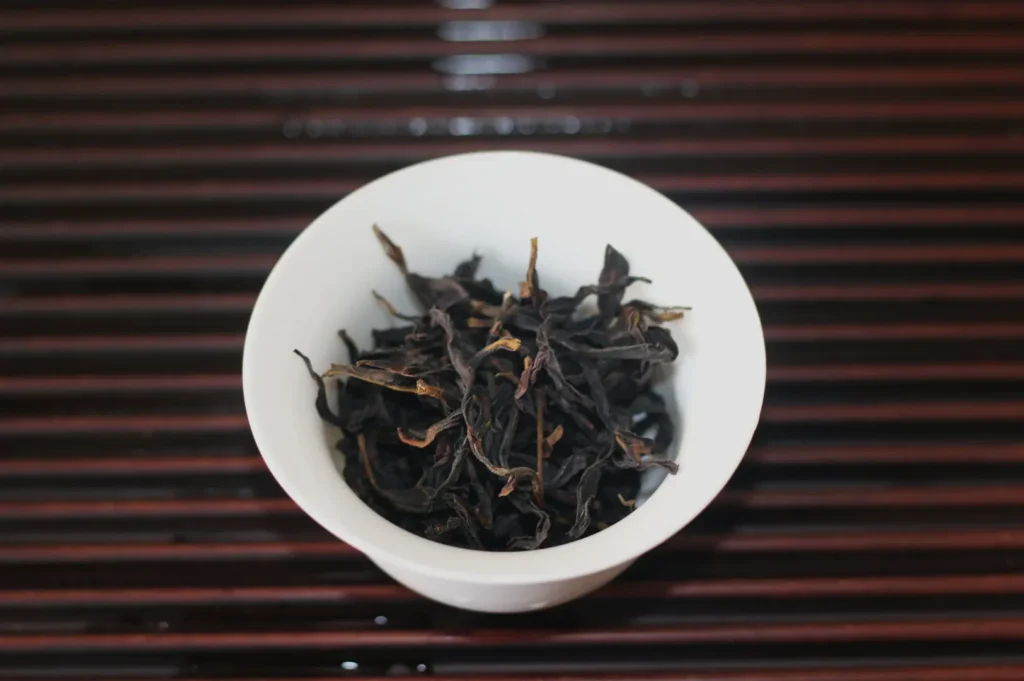 A white bowl filled with loose, dark dried tea leaves sits on a black-and-red striped surface, creating a simple, quiet still life.