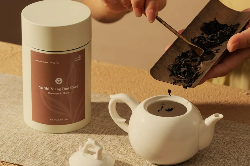 Hands prepare traditional tea, scooping dark loose leaves from a tray into a white teapot beside a labeled tea tin, in warm, calm light.