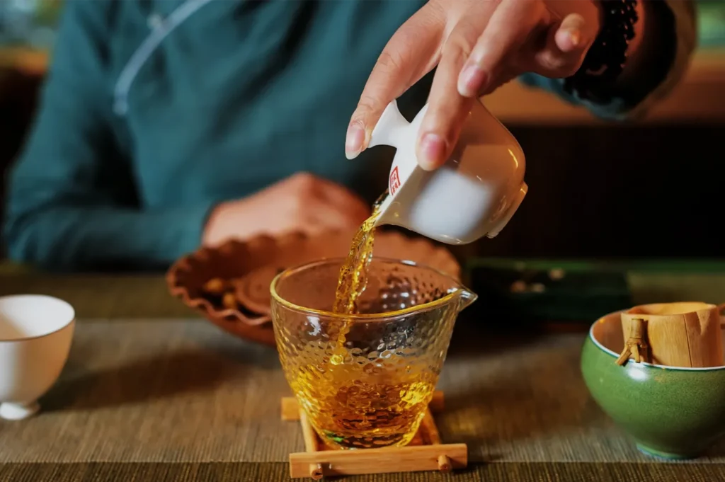 A person pours amber tea from a small white pitcher into a clear glass cup on a wooden table, creating a warm, tranquil tea ceremony scene.