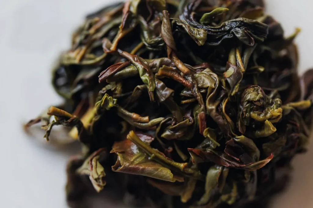 A close-up of loose, twisted tea leaves in dark green, brown, and golden tones, scattered on a white surface; the rich texture feels earthy and rustic.