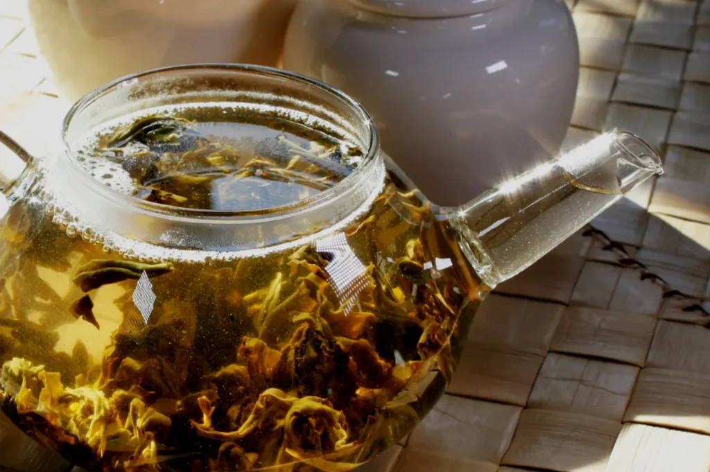 A clear glass teapot holds golden amber tea and unfurling flower petals, glowing in sunlight on a woven bamboo mat; calm, inviting mood.