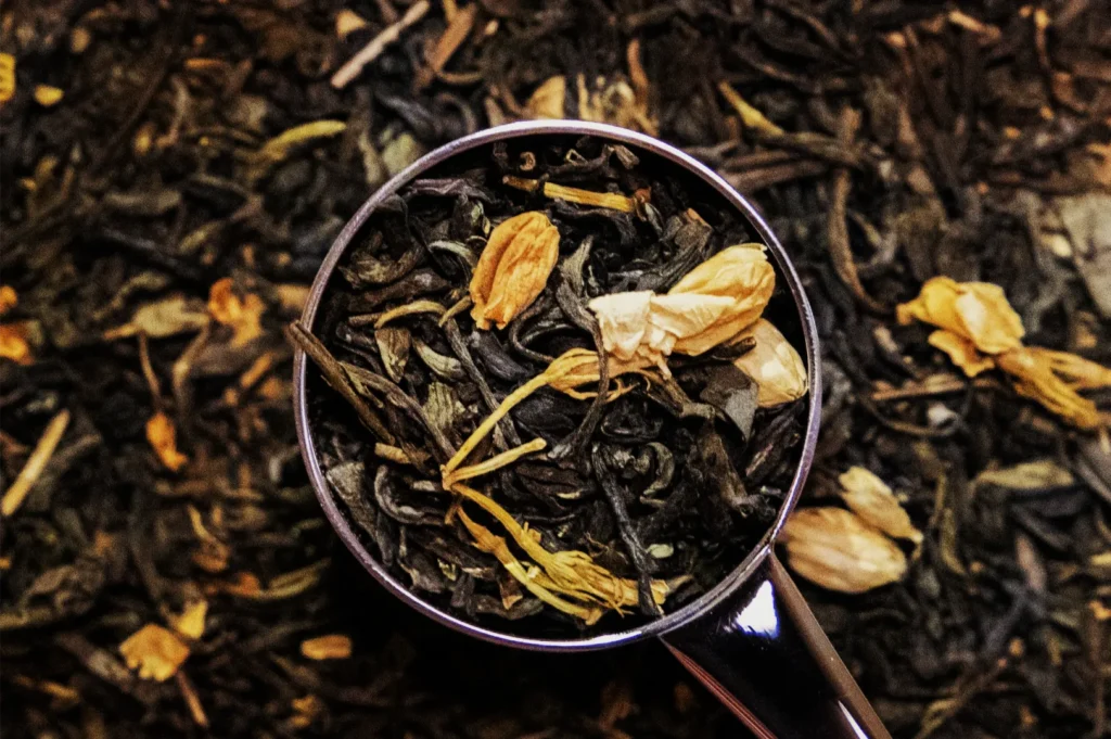 A metal spoon filled with loose black tea leaves and golden dried flower petals rests on a bed of tea, creating a warm, rustic still life.