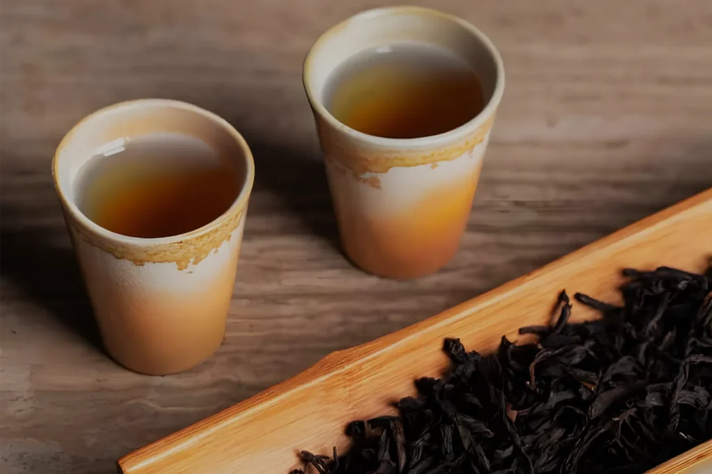 Two cream-colored ceramic cups of amber tea sit on a rustic wooden surface beside loose dark tea leaves, creating a warm, calm mood.