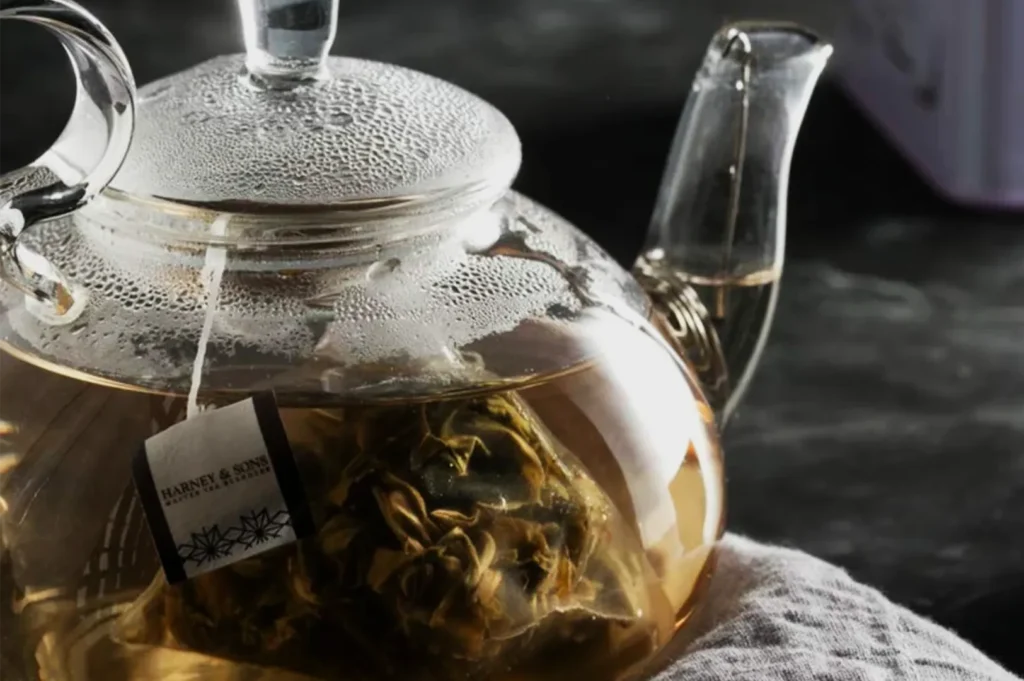 A clear glass teapot steeps blooming herbal tea, with golden liquid and leaves visible through the pot; soft light creates a warm, calming mood.