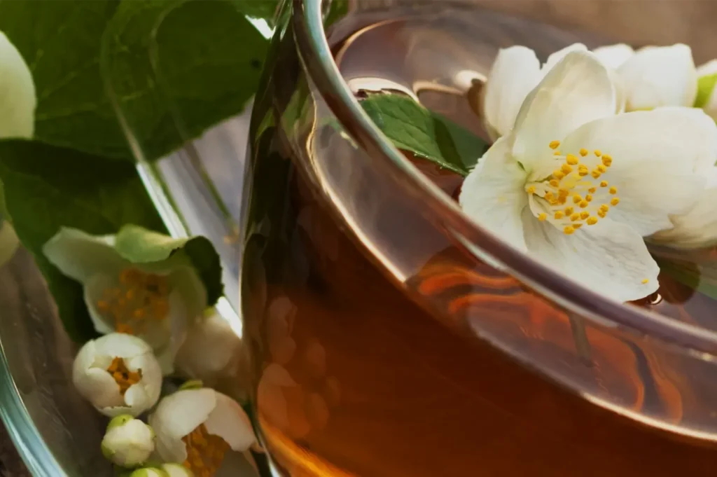 Close-up of a clear glass teacup filled with amber tea and topped with white jasmine blossoms, beside green leaves and buds; serene, natural mood.