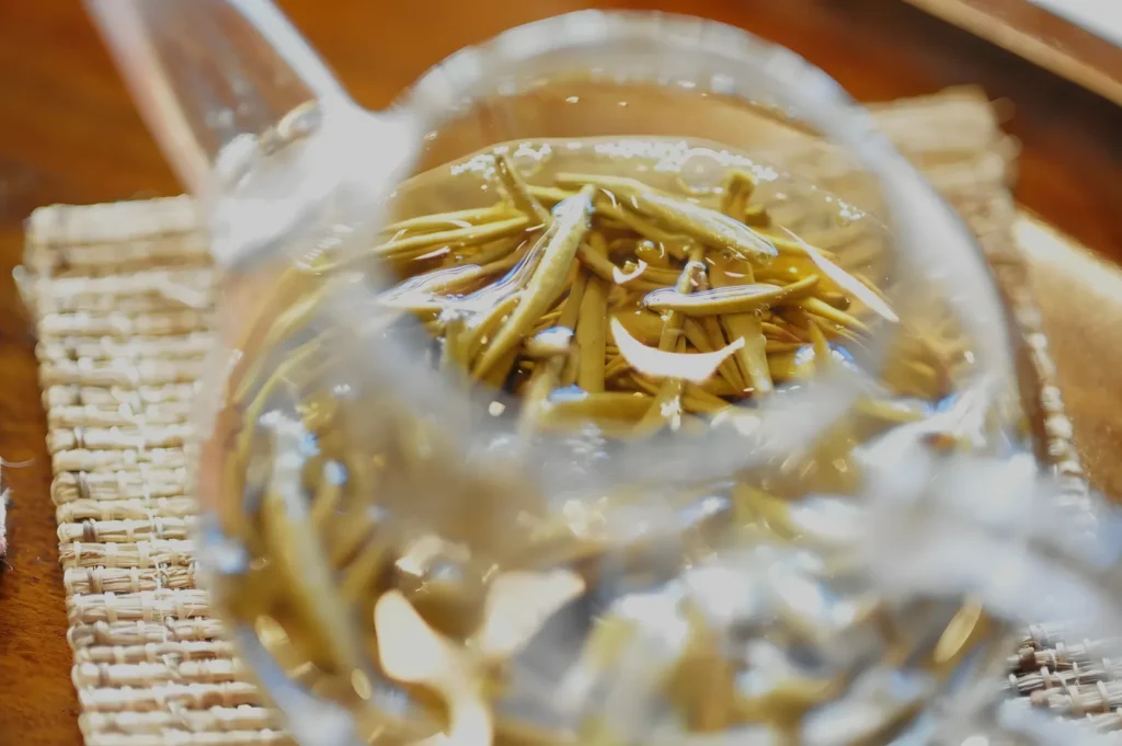 A glass teapot of golden herbal tea with floating rosemary sprigs sits on a woven mat, creating a warm, soothing and inviting mood.