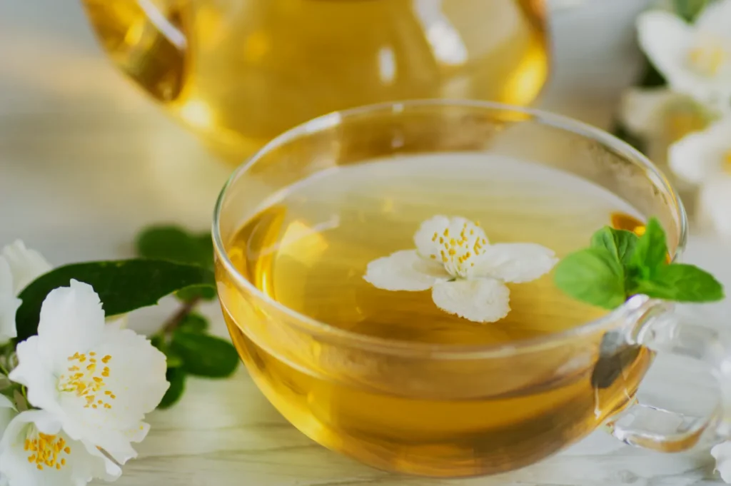 A glass cup of golden herbal tea topped with a white blossom sits among fresh white flowers and green leaves, creating a calm, soothing mood.
