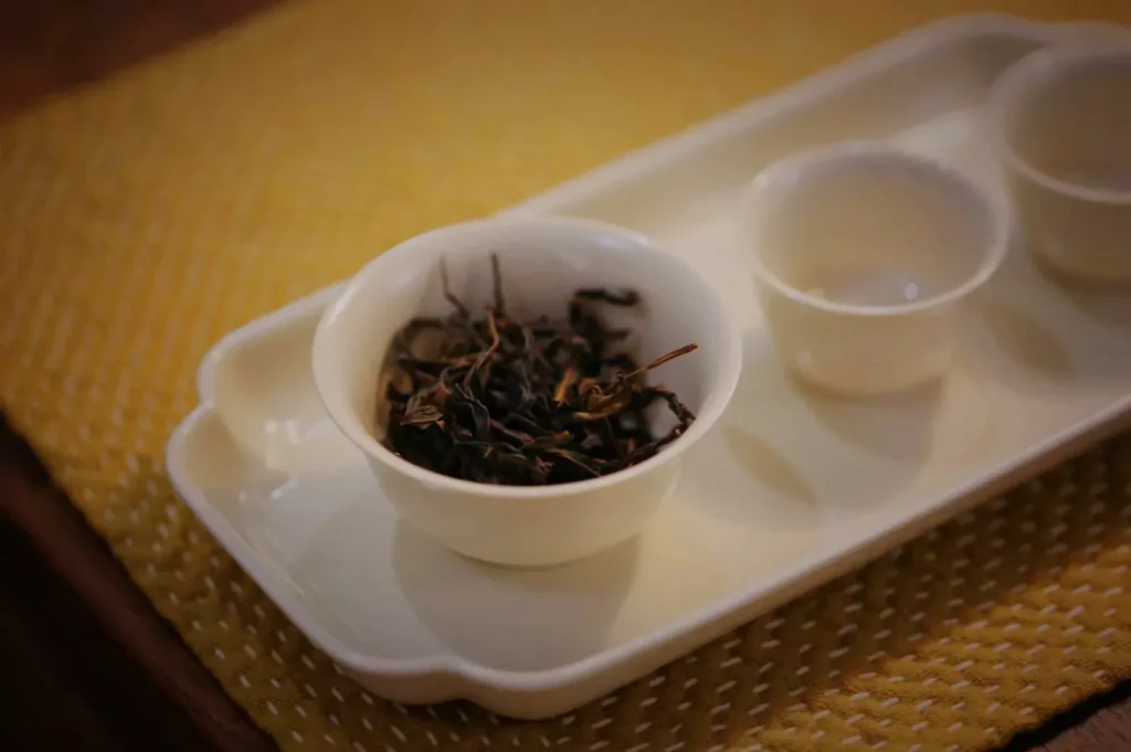 A white tea tray holds a bowl of loose dark tea leaves and small tasting cups on a golden woven mat, creating a warm, calm mood.